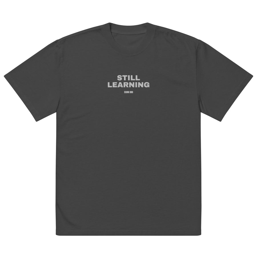 Front mockup of a charcoal tee with light STILL LEARNING lettering centered on the chest.