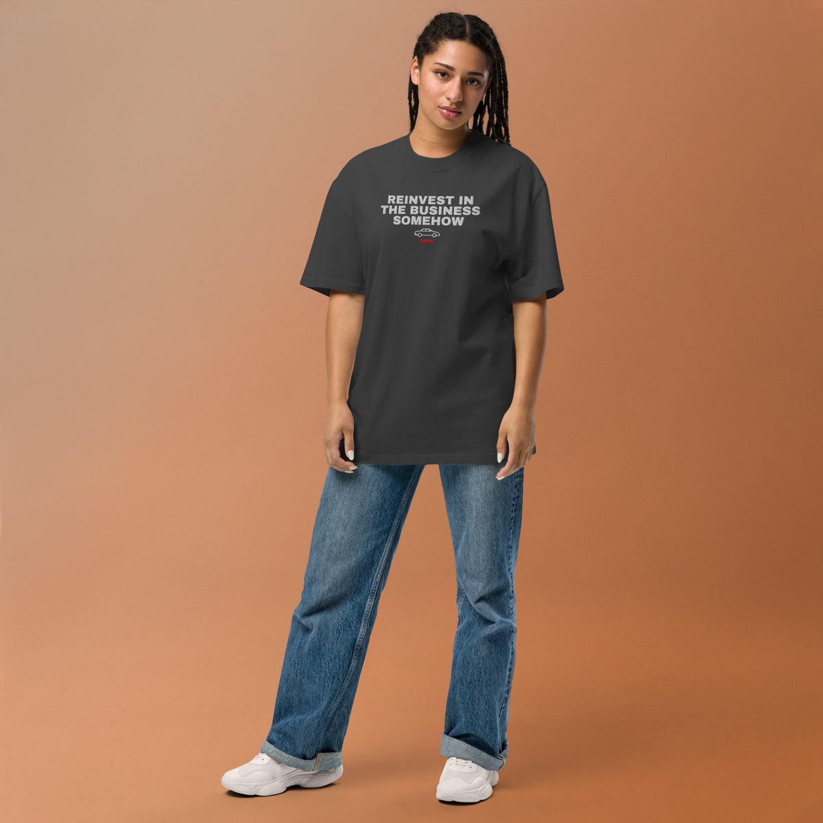 Supplier mockup: model in blue jeans wearing the Faded Black Reinvest in the Business Somehow tee, with white embroidered headline, coupe icon and red ECOM BRO signature on the front.