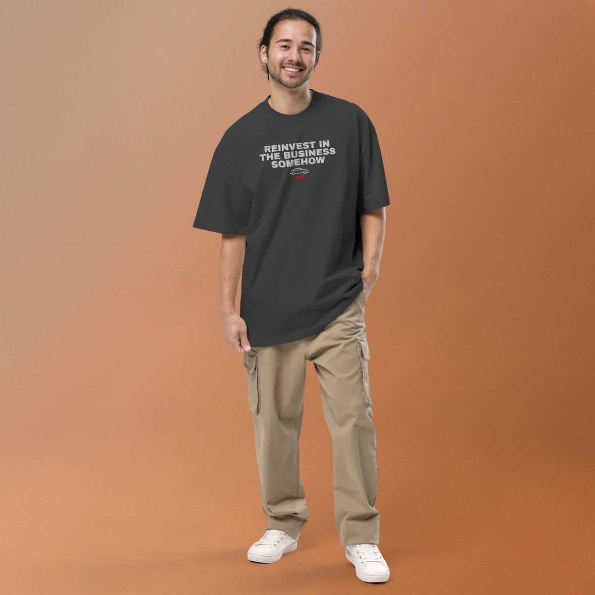 Supplier mockup: model in beige trousers wearing the Faded Black Reinvest in the Business Somehow tee, with the embroidered headline, coupe icon and ECOM BRO signature on the front.