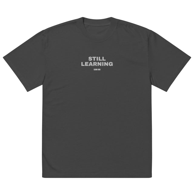 Front mockup of a charcoal tee with light STILL LEARNING lettering centered on the chest.